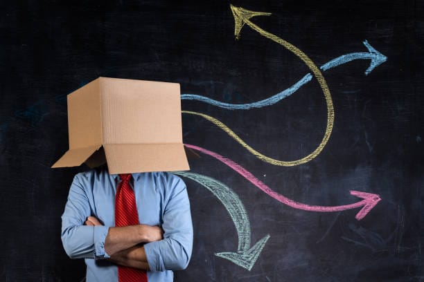 Businessman hiding his head in a cardboard box with crossed arms in front a blackboard showing many multicoloured arrows going in different directions. It is a sing of inhibition.