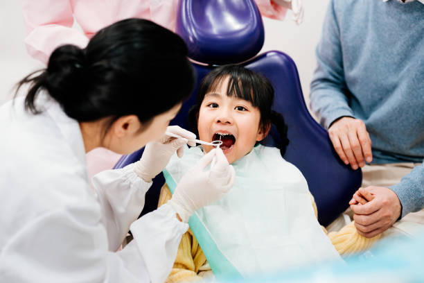 Asian little girl having teeth examined in dental clinic. Female dentist examining her teeth, her father was trying to calm her down. Dental treatment for children.Caries prevention in kids.